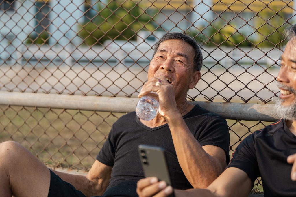 Older man sitting on bleachers drinking a bottled water after working out.